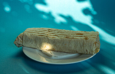 Large shawarma on a white plate with a napkin on a blue background with shadows. Food, healthy fast food, delicious to eat.