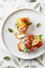 Bruschetta sandwiches with tomatoes, cream cheese, olive oil and basil on white plate on printed tile background close up, top view. Traditional italian antipasti