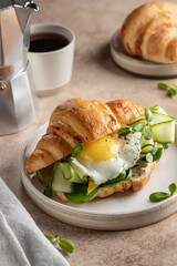 Croissant sandwich with fried egg, cheese, cucumber and micro greens and coffee over beige background. Healthy delicious breakfast