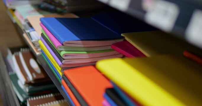 Close-up of stack of multicolored notebooks and exercise books on shelf in shop. Heap of composition books of solid vibrant colors lie for sale in store.