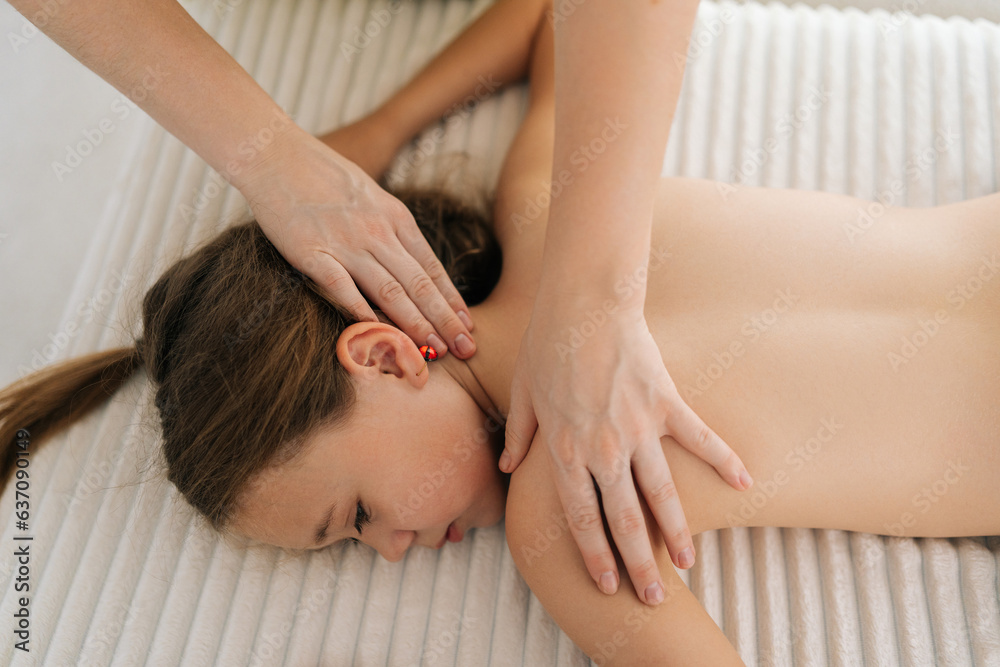 Wall mural Top view of cute little girl having neck, shoulder and back massage by unrecognizable female masseuse lying on massage table. Adorable preteen kid getting physiotherapy from masseuse for body care.