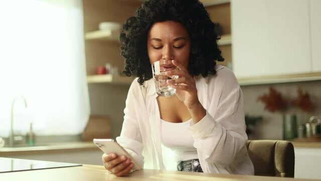 Charming young curly woman drinking clean water from a glass while scrolling browsing on smartphone checking email at home Pretty female keep health balance hydration during day indoors