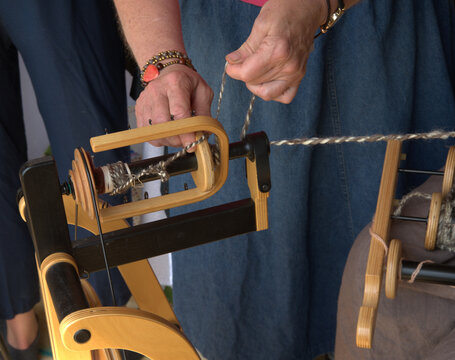 A weaver adjusts the threads on the spindle of a wooden spinning wheel that is driven by foot pedals. 
