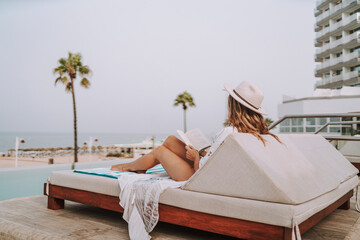 Chica joven en hamaca con smartphone tomando fotografías y leyendo en piscina de hotel de lujo