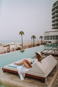 Chica joven en hamaca con smartphone tomando fotografías y leyendo en piscina de hotel de lujo