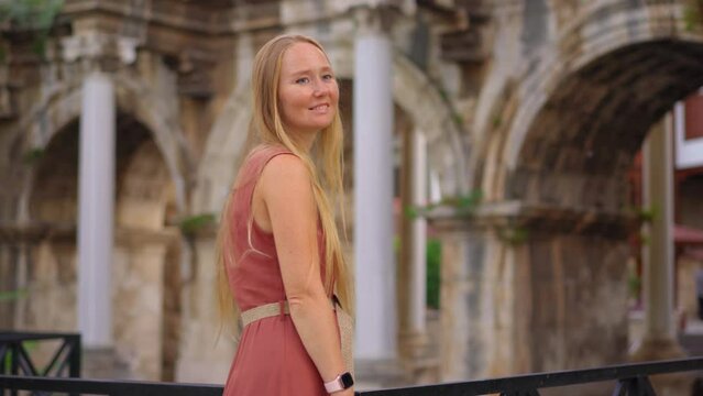 In this captivating video, a young woman tourist is portrayed visiting the renowned tourist attraction in Antalya - Hadrian's Gate. She stands before the majestic archway, adorned with intricate