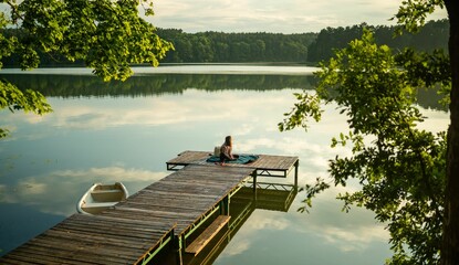 Young female sitting on a wooden dock at a lake, surrounded by tall trees and foliage