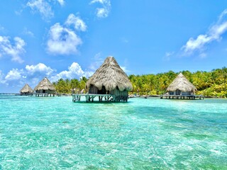 Traditional thatched huts in a tranquil turquoise ocean with palm trees in the background. Belize.