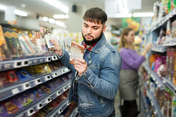 Young bearded guy choosing chocolate on shelves in grocery store