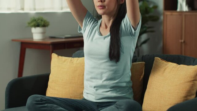 Happy peaceful young Asian woman sit cross-legged eyes closed doing meditation practice on sofa. Relaxing girl meditate felling balance in life improve improve self-conscious at home living room