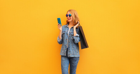 Beautiful happy young woman looking at mobile phone with shopping bag wearing denim jacket on...