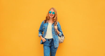 Portrait of stylish modern smiling young woman with backpack wearing denim jacket on yellow studio...