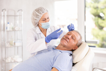 Obraz premium Old male patient lying on clinical chair undergoing face care procedure by injection method