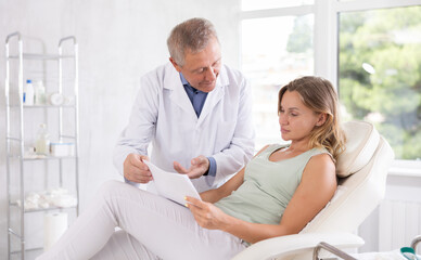 Obraz premium Senior man doctor gives middle-aged female patient sitting in chair medical documents for review before carrying out cosmetic procedure. Medical documentation, questionnaire, patient survey