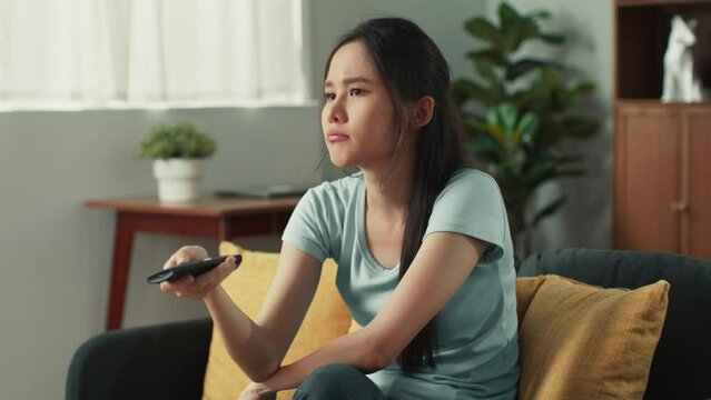 Attractive young asian woman sits on a sofa watching television using remote control changing channel on tv in living room at home. Asian woman having fun during weekend watch movie on tv at home.