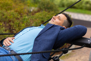 Adult handsome male businessman resting on hammock in park during break at work. Balance between work and personal life. Man in jacket lying on bench on nature