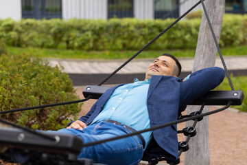 Adult handsome male businessman resting on hammock in park during break at work. Balance between work and personal life. Man in jacket lying on bench on nature