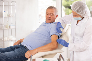 Obraz premium Old male patient lying on clinical chair having arm injection by middle-aged doctor