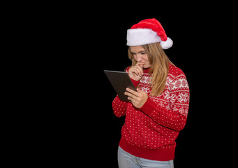 Young blonde blue-eyed girl uses a tablet with a thoughtful face. light-eyed woman in a red christmas sweater and santa hat poses with a table on a black background.