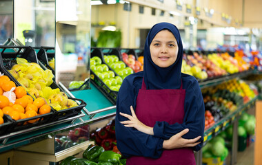 Young woman seller in hijab posing near shelves with vegetables and fruits in grocery store