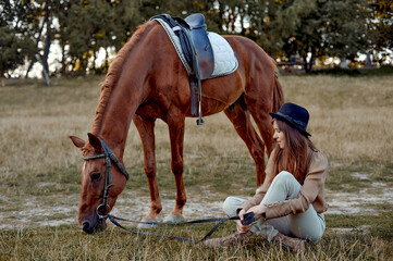 In the countryside, a young woman in black hat sits on grass near a brown horse grazing grass....
