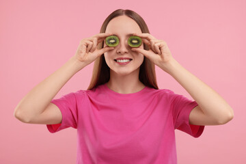 Young woman holding halves of kiwi near her eyes on pink background