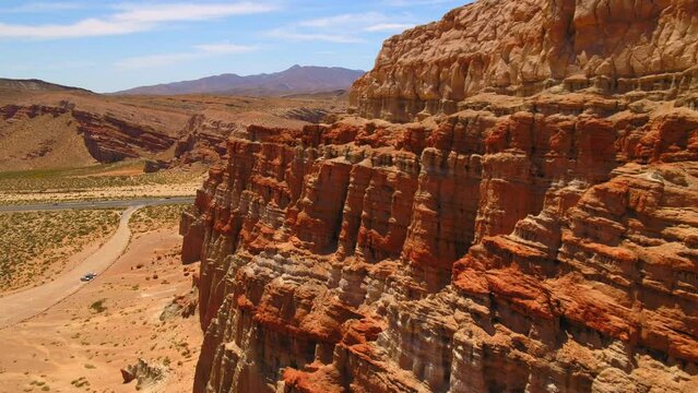 Aerial Forward Shot Of Red Rock Cliffs Near Cars And Mountain Ranges Against Sky During Sunny Day - Red Rock Canyon State Park, California
