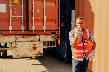 African American male worker standing outdoor work behind truck uses radio to communicate with crews of port delivery drivers to move cargo in logistics container trucks to deliver to customers. - obrazy, fototapety, plakaty
