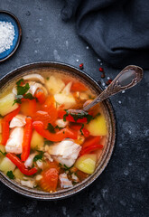 Fish soup with cod, red paprika, potatoes, tomatoes and parsley in soup bowl on black table background, top view