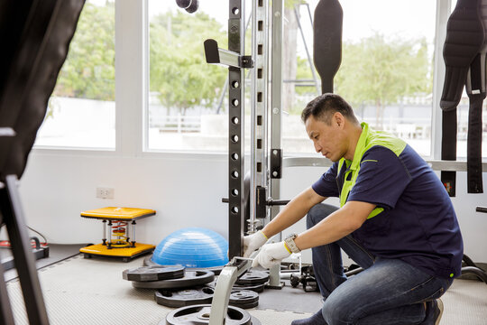 Professional Asian male service worker or fitter checks equipment, maintains and secures fitness equipment in indoor gym, provides safety for users : Skilled technicians repair exercise machines.