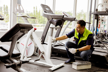 Professional asian male service repair worker or sport fitter using bicycle and treadmill service equipment, supervising and securing fitness equipment in indoor gym.