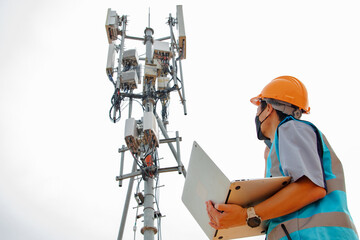 Helmeted asian male engineer works field with telecommunication tower that controls mobile electrical installation using laptop to monitor and maintain 5G networks installed in high rise buildings.
