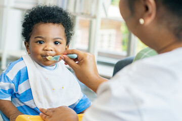 Mother raising take care an African-American Thai son feeds him simple and nutritious lunch : Portrait baby boy who enjoys eating food relishing and gluttonous. - obrazy, fototapety, plakaty
