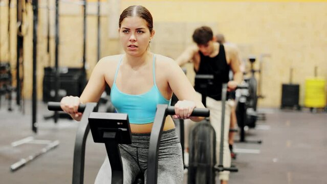 Young motivated fitness woman warming up and pedaling on exercise bike during group CrossFit workout in gym health center
