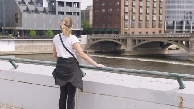 Young woman taking in the view from a bridge over a river in a downtown, urban environment.  Shot in 4K