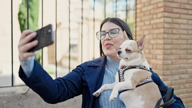 Young hispanic woman with chihuahua dog smiling confident taking a selfie picture at street