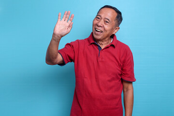 Smiling elderly friendly Asian male waving hands saying goobye or hello isolated on blue background