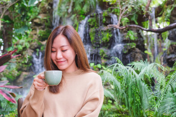 Portrait image of a young woman holding and drinking coffee while sitting in the garden with waterfall