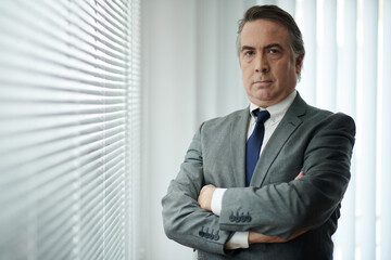 Portrait of serious confident businessman standing at office window with arms crossed