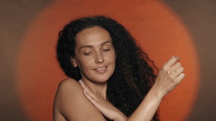 A woman enjoys the softness and silkiness of her skin, touches her shoulder. Portrait of a seminude curly woman in the studio on an orange background with circular light. - obrazy, fototapety, plakaty