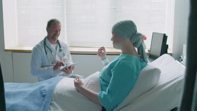 Medium shot of young woman with cancer communicating with doctor while lying on bed in hospitals ward