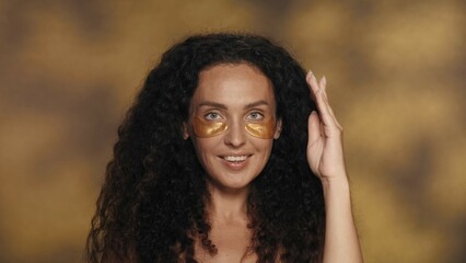 Longhaired curly brunette woman performs cosmetic procedures. Portrait of a seminude woman with golden patches under her eyes in the studio on a yellow background with highlights close up.