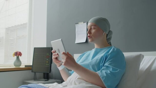 Medium shot of young female cancer patient having video call with her parents while laying on bed in hospitals ward