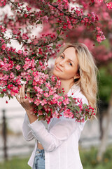 Fototapeta premium Charming long-haired beautiful blonde with perfect skin is standing touching her face with pink flower petals cherry blossoms in the sun