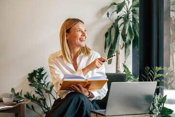 Smiling woman manager working on laptop, making notes in cozy coworkign space interior