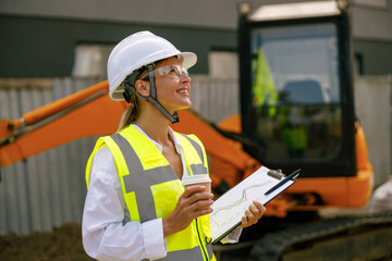 Female engineer in protective helmet drinking coffee standing against tractor on construction site