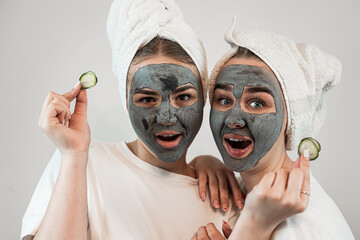 Two girlfriend or sister wear white towel with black clay mask hold pieces of cucumber