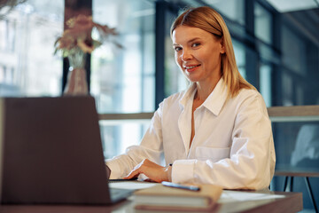 Woman entrepreneur working on laptop while sitting in cozy coworking and looking at camera