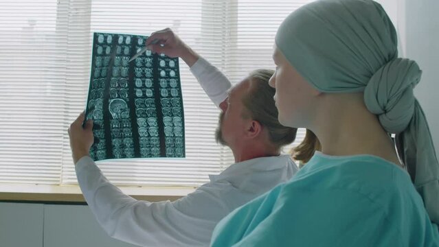 Over the shoulder of patient and doctor talking about brain X-ray image scan while sitting in ward of hospital