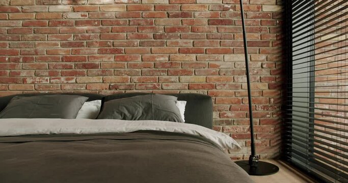Empty comfortable bed with pillows covered gray blanket at modern hotel room with loft interior, brick wall and panoramic window with blinds, slow motion. Stylish bedroom with contemporary design
